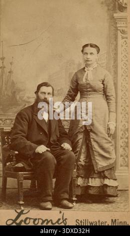 CDV homme est assis dans une chaise portant un costume sombre, et la femme se tient à côté de lui dans une robe formelle avec une agitation. Banque D'Images