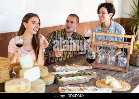 Un homme et deux femmes appréciant la dégustation de fromage tout en tenant des verres avec du vin rouge sur une table en bois rustique dans le restaurant Banque D'Images