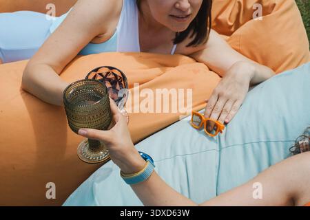 Deux amies se relaxant sur des poufs orange et bleus à l'extérieur tenant des boissons. Concept de loisirs d'été, d'amitié et de vacances par une journée ensoleillée. Banque D'Images