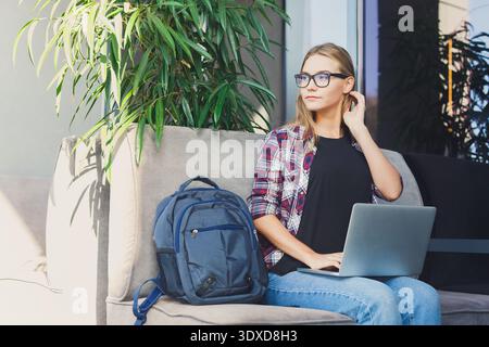 Jeune femme travaille sur ordinateur portable dans un cadre de café moderne entouré de plantes et de lumière vive Banque D'Images