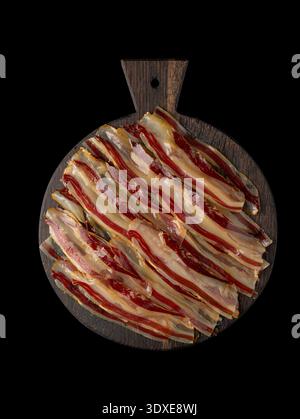 Tranches de bacon crues striées sur une planche de bois foncé. Isolé sur noir, vue de dessus. Copier l'espace. Banque D'Images
