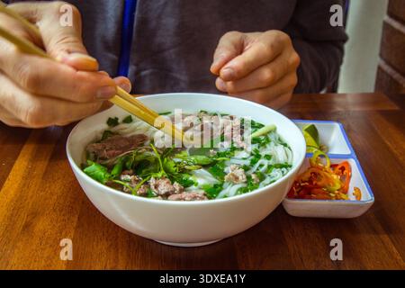 Un homme mange la soupe traditionnelle de bœuf vietnamien Pho Bo avec des baguettes. Nourriture de rue. Manger un bol de pho avec des nouilles et du bœuf. L'homme tient des baguettes Banque D'Images