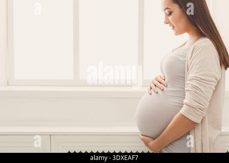 Femme enceinte pose près d'une fenêtre avec ses mains reposant sur son ventre dans un cadre calme Banque D'Images