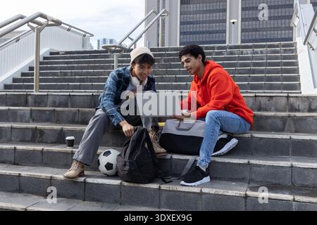 Divers étudiants adolescents assis dans les escaliers à l'aide d'un ordinateur portable argenté, écouteurs, ballon de football à proximité Banque D'Images