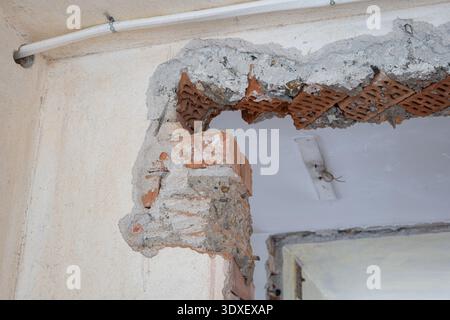 Gros plan d'un coin de mur en brique rouge et béton grossièrement brisé lors d'une rénovation de maison, avec un conduit électrique exposé et du plâtre brut f Banque D'Images