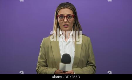 Femme journaliste tenant un microphone avec sourcil sillonné, regardant sur le côté, debout dans le studio avec toile de fond violette ; concerne la mise à jour des nouvelles. Banque D'Images
