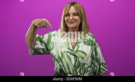 Femme d'âge moyen avec des cheveux blonds et des lunettes pointe doigt vers le bas dans le studio portant une chemise verte de feuille tropicale et souriante ; invitation joyeuse. Banque D'Images