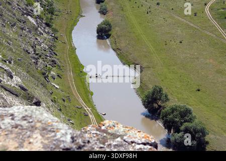 Vue aérienne d'une rivière tranquille et sinueuse depuis une falaise. Une jetée en bois rustique se trouve dans l'eau boueuse, flanquée d'une pente rocheuse et d'un gre Banque D'Images