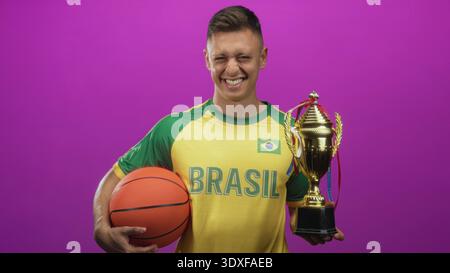 L'homme tient le basket-ball et le trophée et sourit tout en portant le maillot brésilien avec patch drapeau en studio, regardant fier et détendu ; joie de la victoire. Banque D'Images