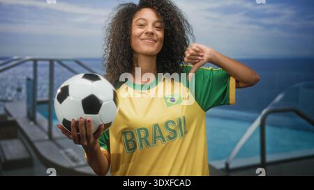 Femme tenant le ballon de football et donnant les pouces vers le bas sur la terrasse de la piscine portant le maillot du brésil et souriant ; fierté. Banque D'Images