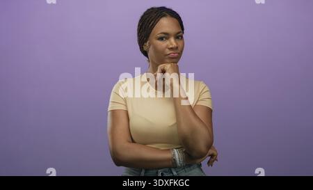 Femme avec la main sur le menton et les bras pliés debout dans le studio à côté du mur violet portant un tee-shirt beige et un Jean denim ; pensée contemplative. Banque D'Images