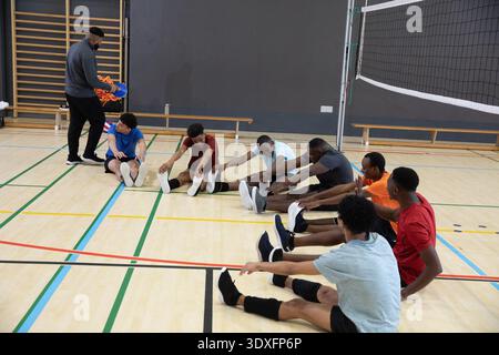 Divers coéquipiers masculins en équipement assis sur le terrain près du filet de volley-ball s'étirant comme entraîneur tenant des cônes. Athlètes, entraînement, gymnase, travail d'équipe, concentration, marqueurs, équipement || autorisation du modèle Banque D'Images