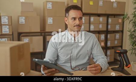 L'homme tape sur l'écran de la tablette avec la main visible dans le bâtiment parmi les boîtes empilées, le lecteur de codes-barres et les étagères ; l'accent est mis sur l'efficacité. Banque D'Images