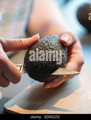 Gros plan des mains coupant un avocat mûr avec un couteau sur une planche de bois. La lumière naturelle souligne la texture rugueuse de la peau et une préparation soignée. Banque D'Images