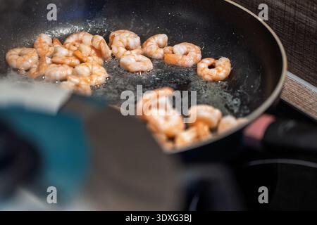 Les crevettes fraîches font frire dans de l'huile chaude à l'intérieur d'une poêle noire sur une cuisinière. La vapeur monte pendant que l'huile bouillonne autour des fruits de mer. Banque D'Images
