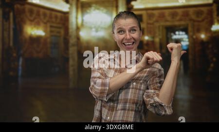 Femme en chemise à carreaux pointe les pouces sur le côté avec un clin d'œil à l'intérieur d'un bâtiment de musée orné ; curiosité ludique. Banque D'Images