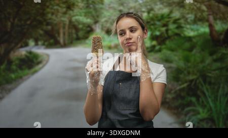 Femme tenant de la crème glacée et portant un tablier avec des gants jetables, doigt à la joue sur une route forestière ; doute de tentation. Banque D'Images