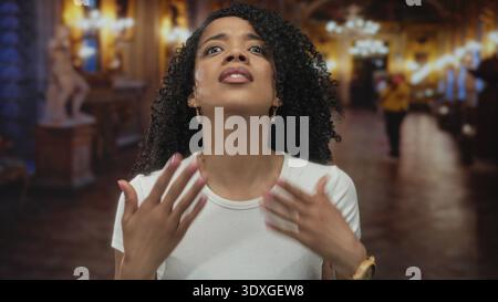 Femme ajuste les cheveux bouclés et soulève le regard sous le grand lustre dans le bâtiment orné du musée ; admiration d'art. Banque D'Images