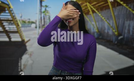 Jeune femme latine couvrant les yeux avec la main et souriant tout en se tenant dans la rue sous la structure métallique ; ludique. Banque D'Images