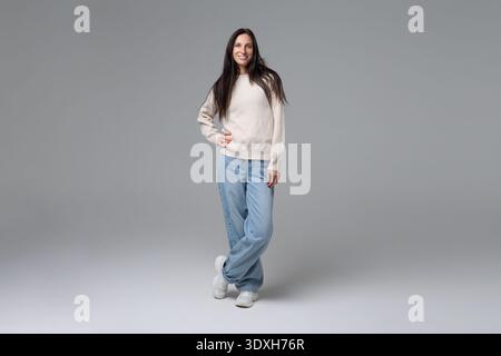Femme heureuse posant en pull blanc et Jean bleu sur fond gris pleine longueur Banque D'Images