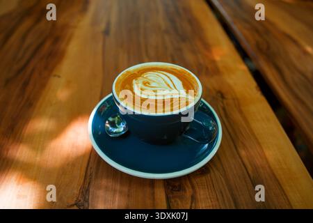 Australie, Queensland, Brisbane. Latte artistique en forme de cœur dans une tasse en céramique bleue avec soucoupe et cuillère, sur une table en bois magnifiquement granulaire Banque D'Images