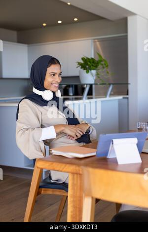 Femme portant un foulard sombre avec des écouteurs assis à la table à manger dans la cuisine moderne à l'aide d'une tablette Banque D'Images