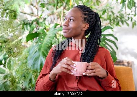 Rêvant femme afro-américaine tenant la tasse reposant sur le fauteuil entourent les plantes d'intérieur regardant de côté. Banque D'Images