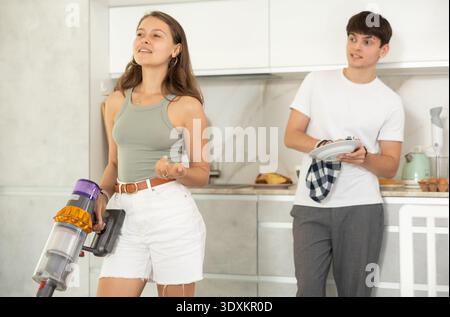 Jeune femme aspirant dans la cuisine tout en communiquant avec son petit ami séchant la vaisselle avec une serviette Banque D'Images
