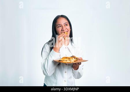 Une joyeuse femme indonésienne dans une chemise blanche appréciant un morceau de Martabak Manis (crêpe sucrée) d'une assiette en bois sur un fond blanc. Banque D'Images