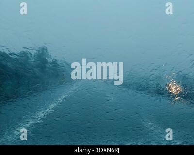 Vue abstraite de la route pluvieuse à travers le pare-brise mouillé de voiture lors d'une journée de pluie lourde | lumières de route floues et gouttelettes d'eau sur fond de texture de verre pour Banque D'Images
