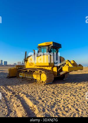 Un bulldozer jaune se tient sur le sable à une plage tôt le matin. L'horizon de la ville peut être vu au loin sous un ciel bleu. Banque D'Images