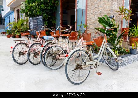 Vélos rustiques alignés à côté d'un café confortable. Les cadres roses et blancs reposent sur du sable mou. Les plantes en pot ajoutent du charme à la scène. Les paniers en cuivre tiennent essentiel Banque D'Images