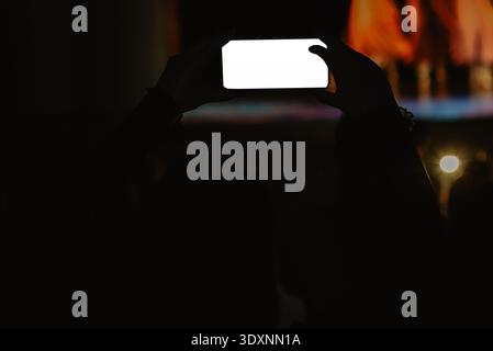 Silhouette d'une personne tenant un smartphone pour enregistrer un concert live ou une performance sur scène la nuit. L'écran lumineux contraste avec la foule sombre, capturant Banque D'Images