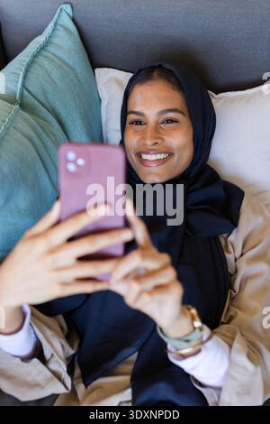 Femme couchée sur un canapé portant un foulard noir et un cardigan beige clair, tenant un smartphone mauve Banque D'Images