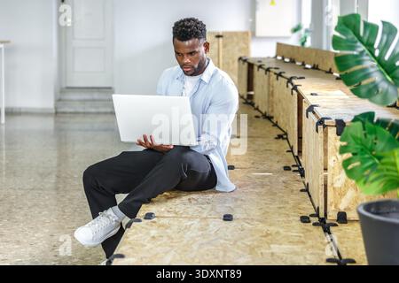 Un jeune homme est assis sur un banc en bois, concentré sur son ordinateur portable dans un espace de coworking lumineux et aéré. La lumière naturelle remplit la pièce tandis que les plantes d'intérieur ajoutent un Banque D'Images