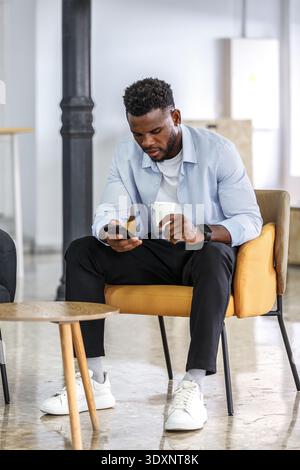 Un homme est assis dans une chaise élégante dans un bureau moderne, concentré sur son smartphone tout en sirotant un café dans une tasse. L'espace dispose d'un décor contemporain et d'un Banque D'Images