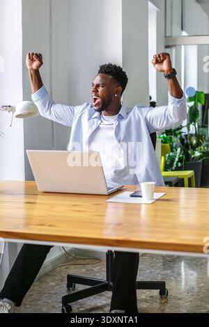 Un homme avec une expression joyeuse lève les bras en célébration tout en étant assis à un bureau. Il travaille dans un bureau lumineux entouré de plantes, un ordinateur portable ouvert Banque D'Images