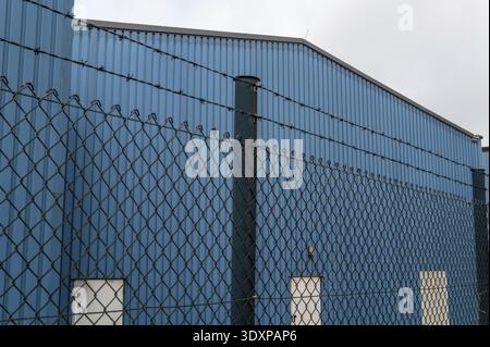 Bâtiment en métal ondulé bleu avec une clôture de maillon de chaîne de sécurité surmontée de fil barbelé Banque D'Images