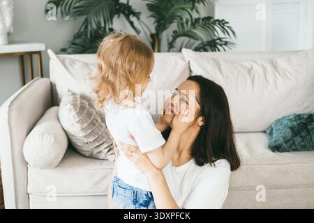 Une mère embrasse doucement sa fille de quatre ans. Fille et mère à la maison sur le sol près du canapé. Le bébé touche doucement le visage de sa mère avec ses paumes. Maman et fille de race mixte. || autorisation du modèle Banque D'Images