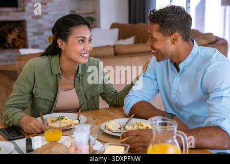 Couple diversifié atteignant et souriant tout en partageant le petit déjeuner à la maison autour d'une table à manger en bois. Intimité, domestique, confortable, rustique, connexion, bonheur, conversation || modèle libéré Banque D'Images