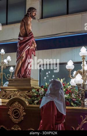 Statue de Jésus-Christ sur un flotteur doré orné lors d'une procession de la semaine Sainte avec un pénitent à capuchon au premier plan, Espagne. Banque D'Images