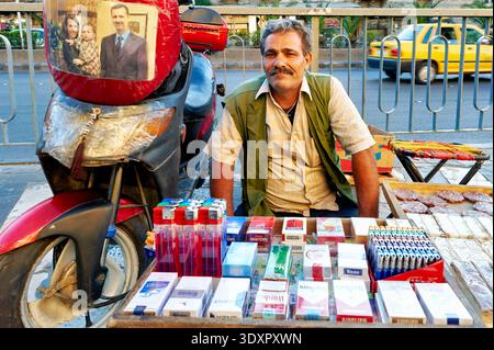 Un homme est assis à une table avec des cigarettes, à côté de sa moto, dans une rue de Damas, en Syrie. Les gens passent en arrière-plan. Banque D'Images