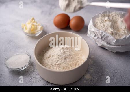 gros plan des ingrédients de cuisson sur un comptoir gris texturé : un bol beige rempli de farine, deux œufs bruns, du beurre et du sucre dans des bols en verre, une croupe Banque D'Images