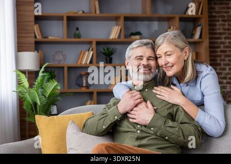 Couple plus âgé assis sur un canapé dans leur salon, embrassant et souriant, représentant des concepts d'amour, de partenariat, de bonheur et d'un style de vie confortable Banque D'Images