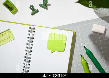Carnet à spirale ouvert avec une note adhésive à bulles de parole verte sur un tapis de découpe gris. Copyspace Banque D'Images