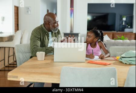 Senior afro-américain homme et enfant fille afro-américaine enseignant à table avec ordinateur portable, notes Banque D'Images