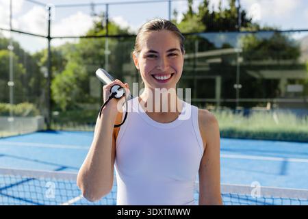 Femme debout sur le court bleu portant le haut athlétique blanc tenant la raquette sur l'épaule souriant Banque D'Images