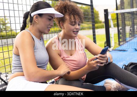 Amies afro-américaines assises sur la cour bleue en vêtements de sport avec visière vérifiant smartphone Banque D'Images