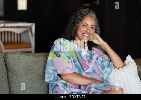 Femme afro-américaine senior assise sur un canapé vert à la maison portant un caftan coloré et souriant Banque D'Images