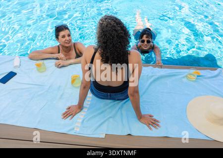 Diverses amies féminines discutant, assis sur la serviette de terrasse de la piscine dans des maillots de bain avec des boissons et téléphone Banque D'Images